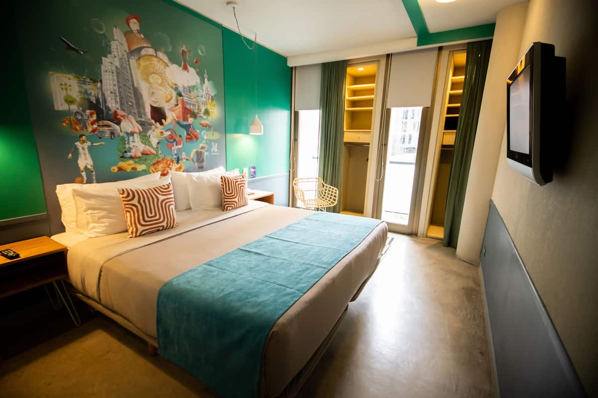 Habitación de hotel con una cama grande, un mural en la pared, un televisor montado y una ventana con cortinas.