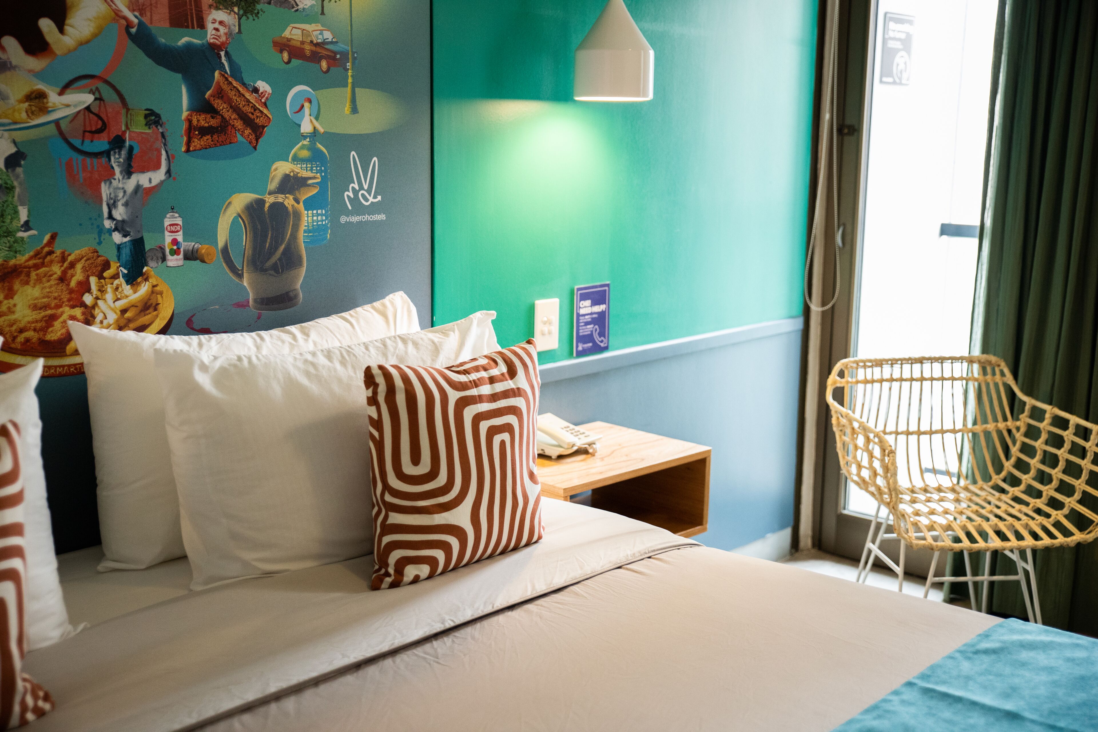 Habitación de hotel con cama, almohadas, un mural en la pared, una mesita con lámpara y una silla de mimbre.