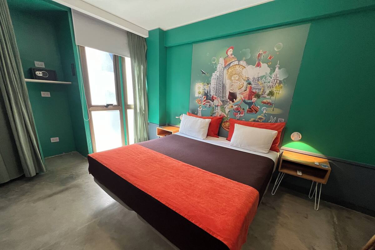 Una habitación de hotel con una cama grande, paredes color aguamarina y un mural colorido en la pared.