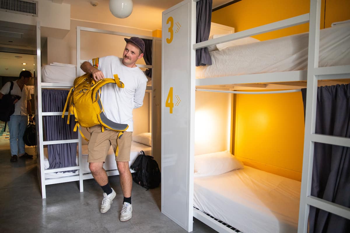 Habitación con literas, los números 3 y 4 en la pared, una persona cargando una mochila y otra al fondo.