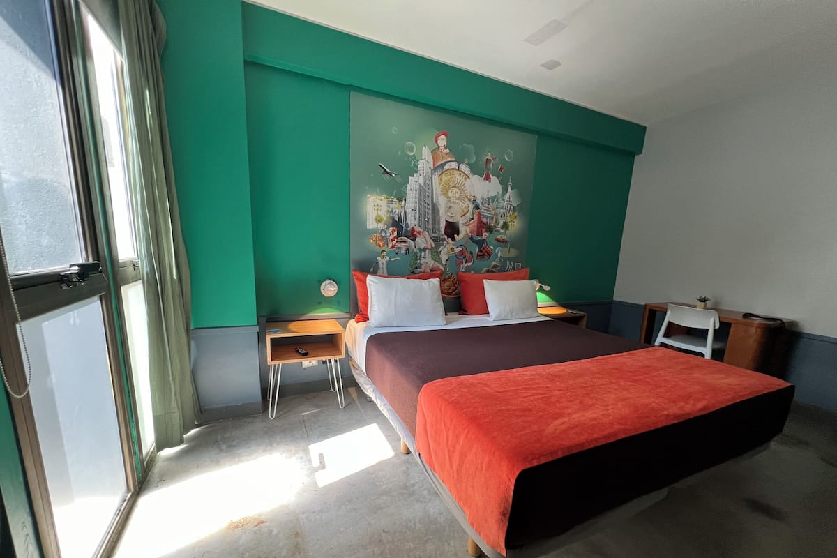 Habitación de hotel con una cama grande, un escritorio, una silla y un mural colorido en la pared.