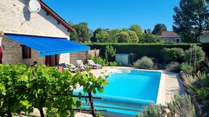 Ein Swimmingpool mit Liegestühlen und einem blauen Sonnensegel, umgeben von Grünflächen und einem Steinhaus.