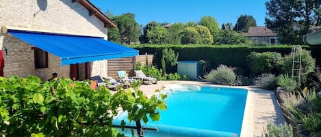 Una piscina con sillas de descanso y un toldo azul, rodeada de vegetación y un edificio de piedra.