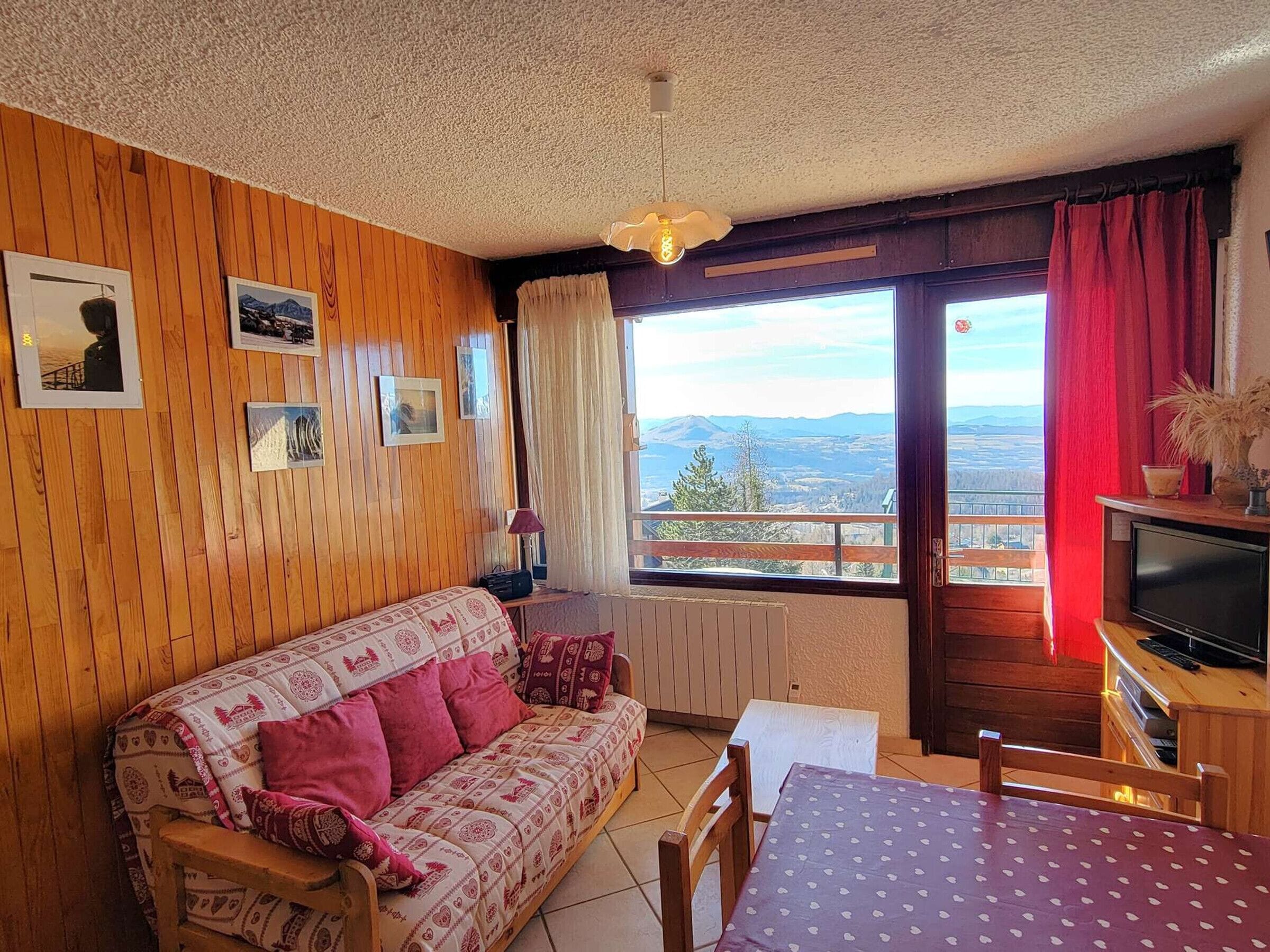 A cozy room with a wooden wall, a sofa, a table, a TV, and a window with a view of the mountains.
