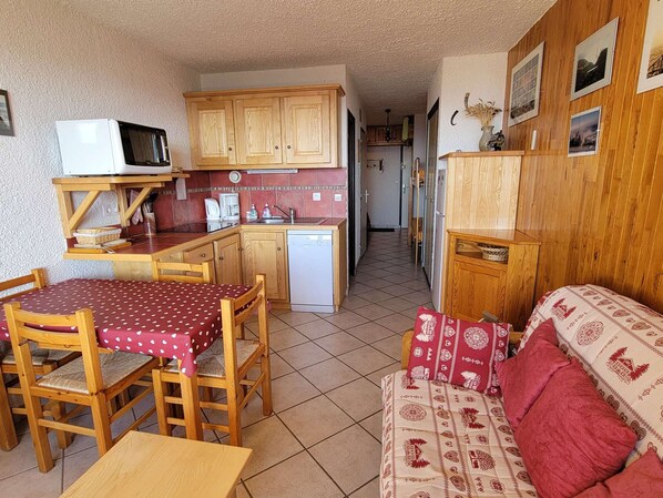 A compact kitchen with wooden cabinets, a microwave, and a dishwasher. There is a dining area with a table and chairs, and a living area with a sofa. - Mountain Studio for 4 with Balcony in Saint-Michel-de-Chaillol (Saint-Michel-de-Chaillol)