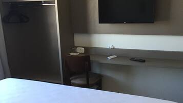 Habitación de hotel con cama, armario, escritorio, silla, televisor y estante con control remoto.