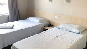 A hotel room with two single beds, a bedside table with a phone, a window with curtains, and an air conditioning unit on the wall.