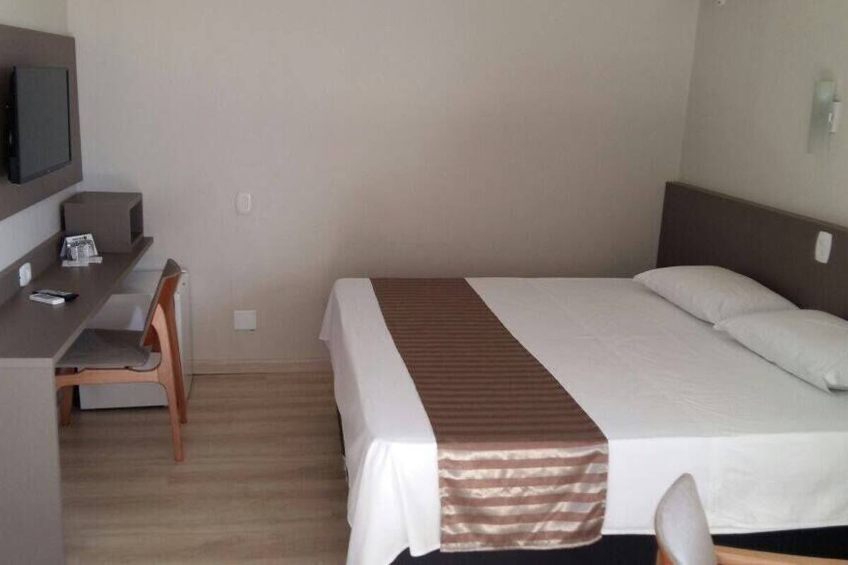 A hotel room with a bed, a desk with a chair, a TV mounted on the wall, and a bedside table.