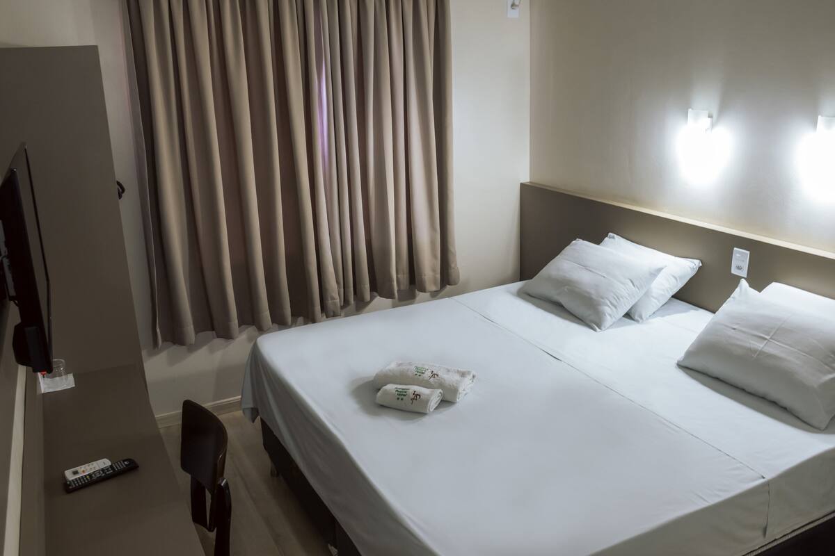 A hotel room with a bed, white pillows, a rolled towel, a remote control, and a television on a stand.