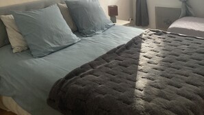 A bedroom with a bed, pillows, a blanket, a wall-mounted lamp, a bedside table, and a window with curtains.