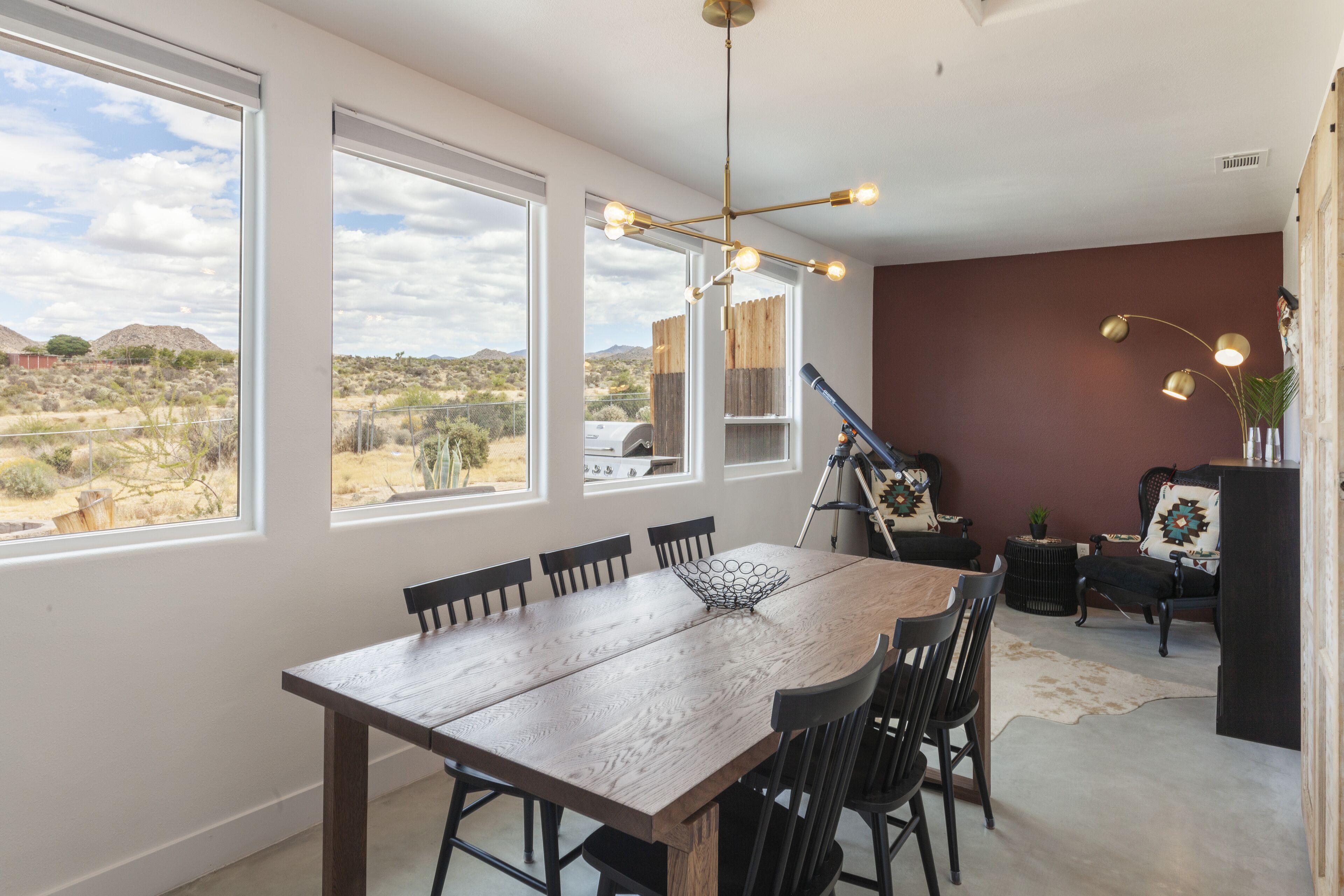 A dining area with a wooden table and chairs, large windows with a desert view, and a modern chandelier.