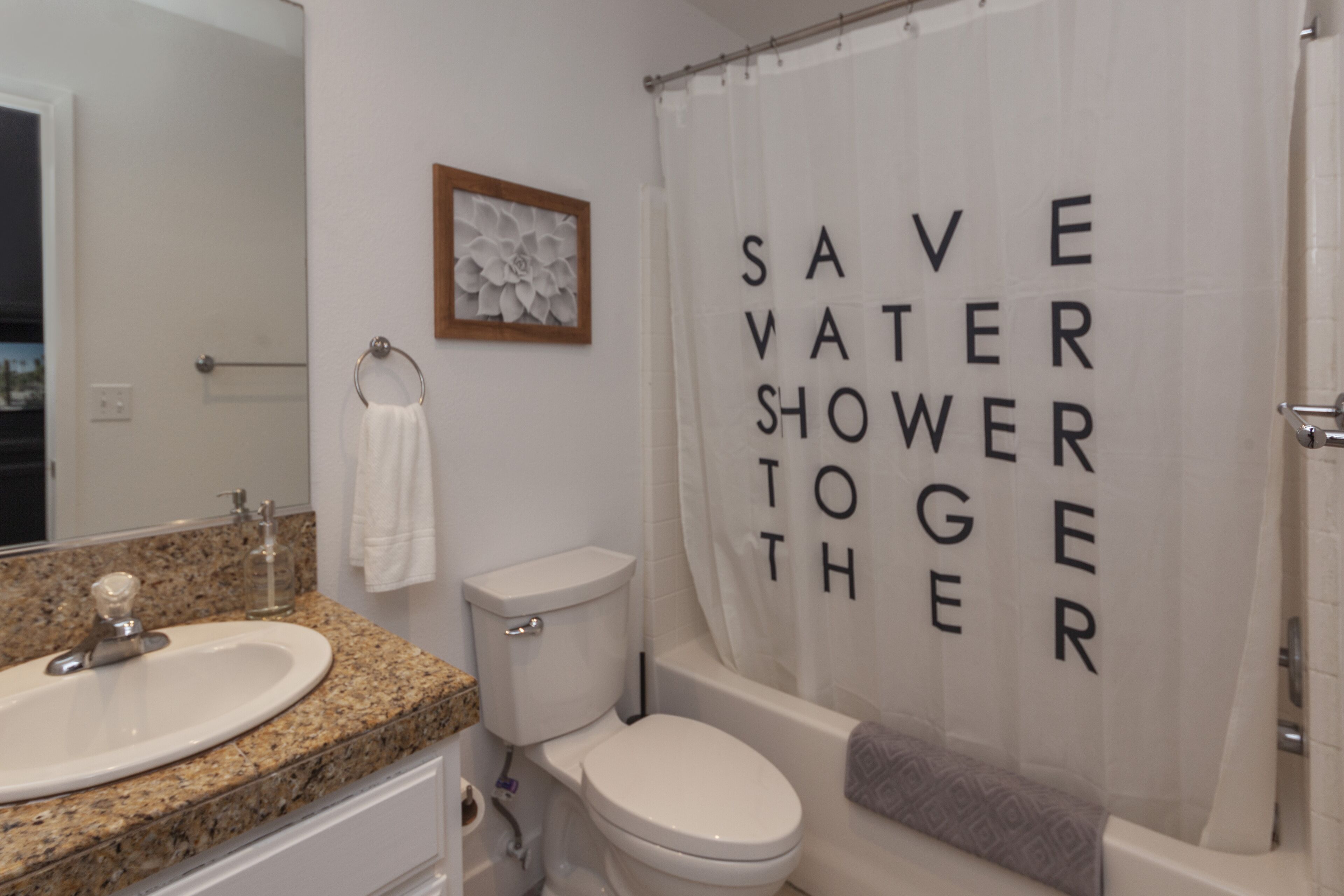 A bathroom with a sink, toilet, and bathtub. There is a shower curtain with the words "SAVE WATER SHOWER TOGETHER" printed on it.