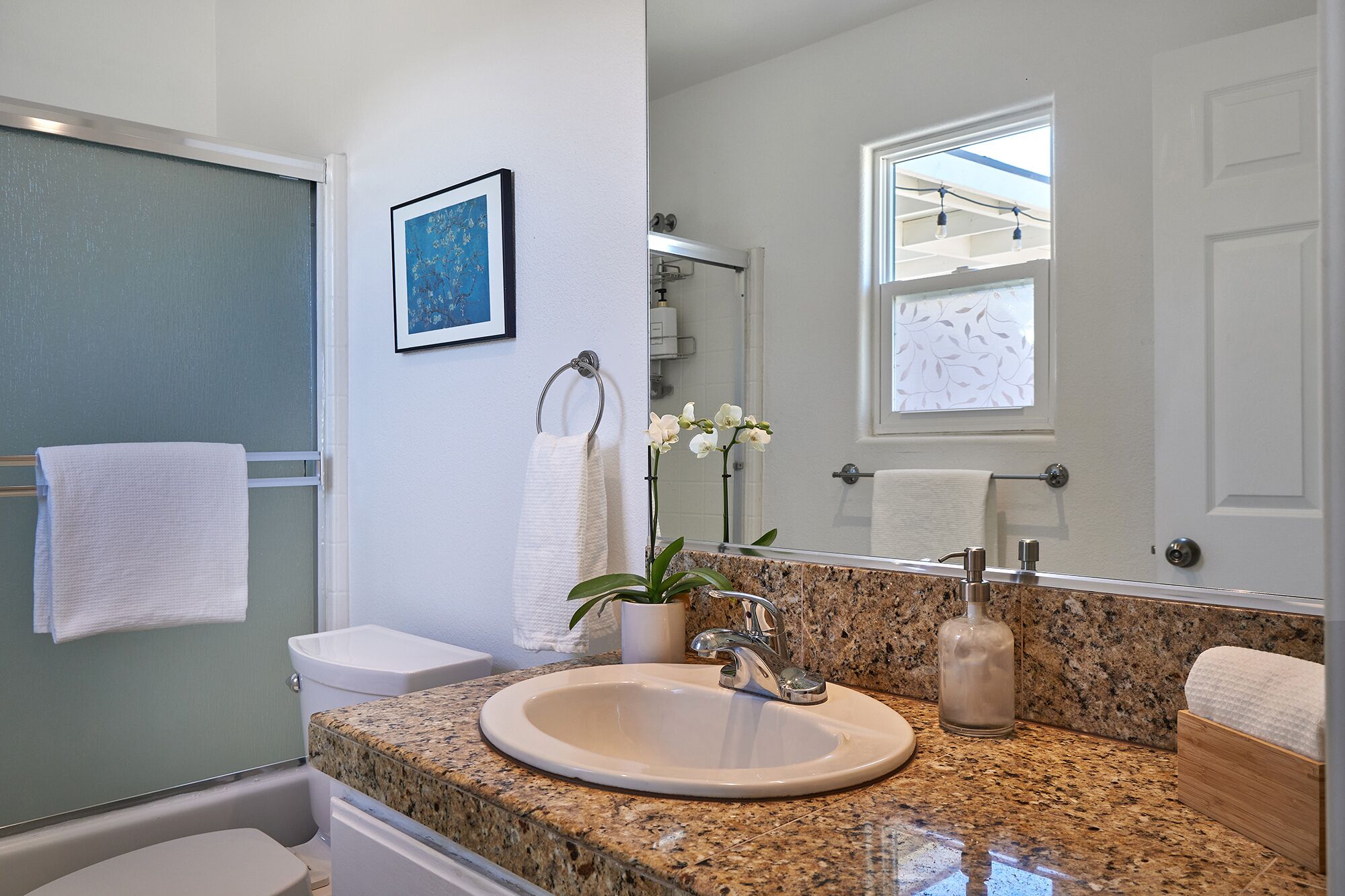 A bathroom with a granite countertop, white sink, and a large mirror. There is a towel rack with white towels and a framed picture on the wall.