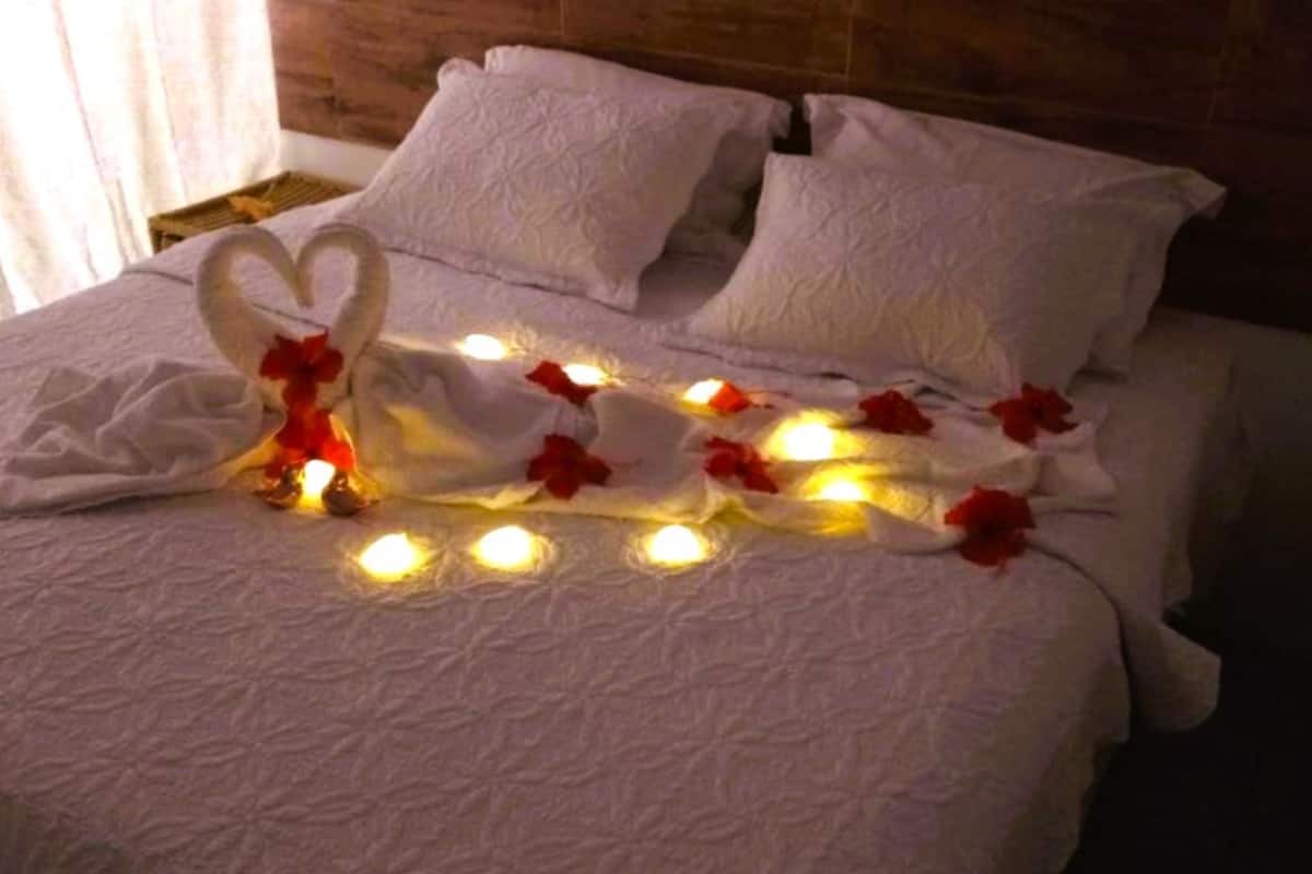 A bed with white linens and pillows, decorated with heart-shaped swans and red roses, and lit by small candles.
