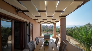 A balcony with a wooden table and wicker chairs, overlooking a swimming pool and a mountainous landscape. - Private Heated pool★Close to the Beach★BBQ & Wifi (Galatas)