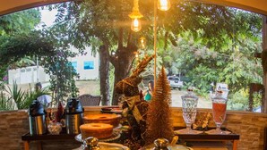 A buffet setup with various food items and a small Christmas tree, set in a room with a view of greenery outside. - Pousada da Leide (Fernando de Noronha)