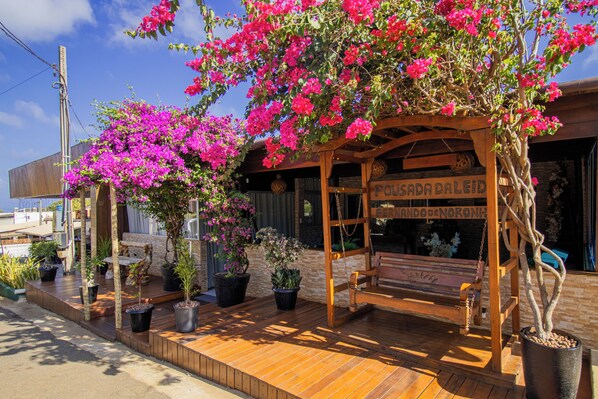 Una pérgola de madera con un banco, rodeada de plantas en macetas y un letrero que dice 'Pavada Daleid'.