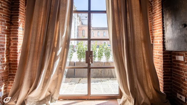 A room with a large window, curtains, and a view of a street with buildings and trees.