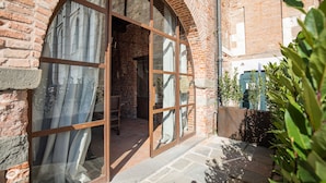 A brick building with a large arched window and a door, leading to an interior space with a tiled floor and a chair.