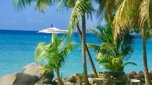 A tropical beach setting with white lounge chairs, a small table, and a palm tree. - Hideaway Apartments Grenada (St. George's)
