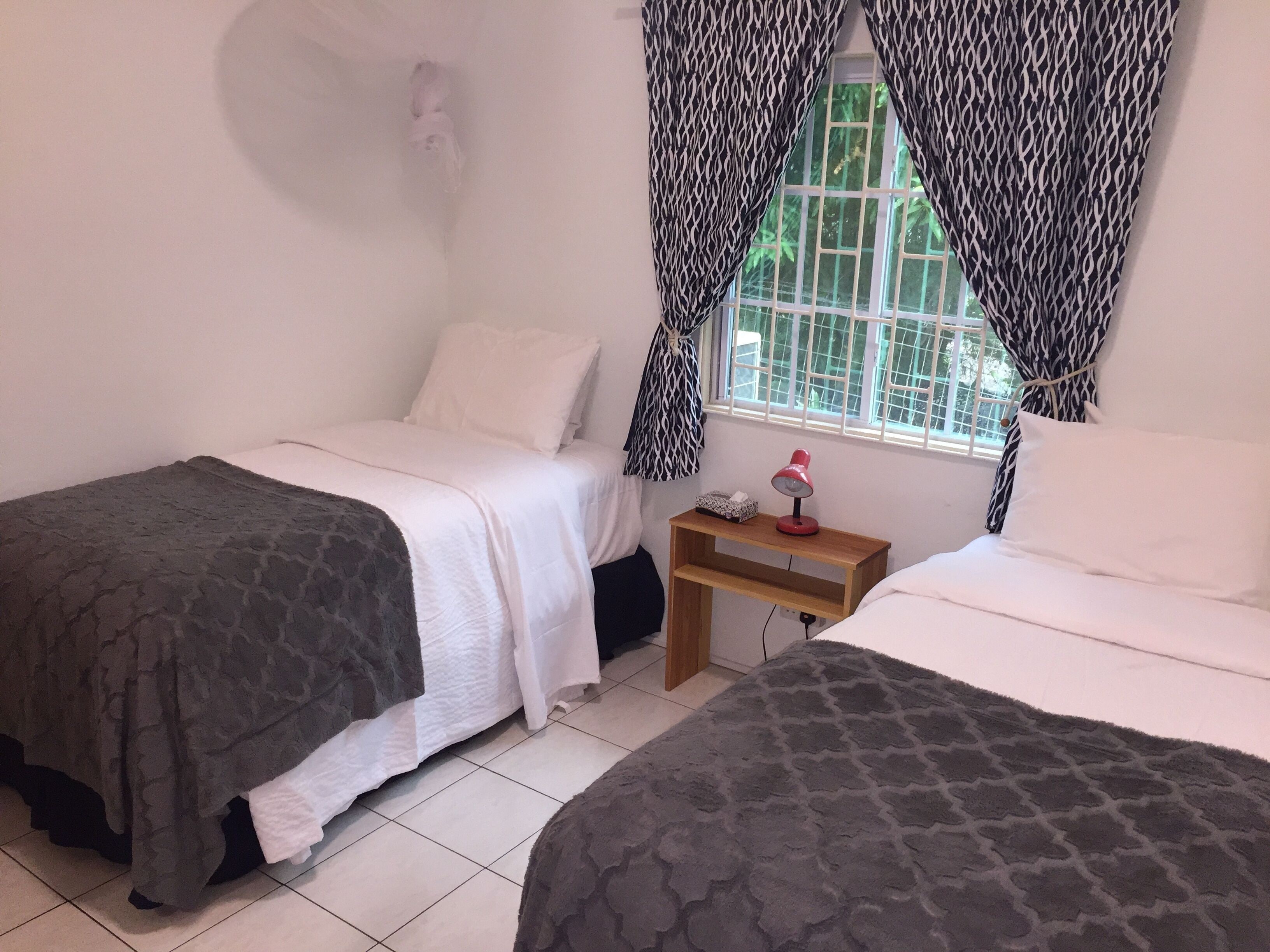 A bedroom with two beds, a bedside table with a lamp, and a window with patterned curtains.