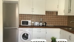 Une cuisine compacte équipée d’un réfrigérateur, d’un four à micro-ondes, d’un grille-pain et d’un lave-linge. On y trouve une table avec des chaises et une plante en pot.