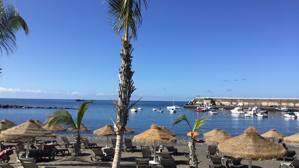 Une vue sur la plage, avec des chaises longues et des parasols en chaume, un ciel bleu dégagé et des bateaux sur l’eau.