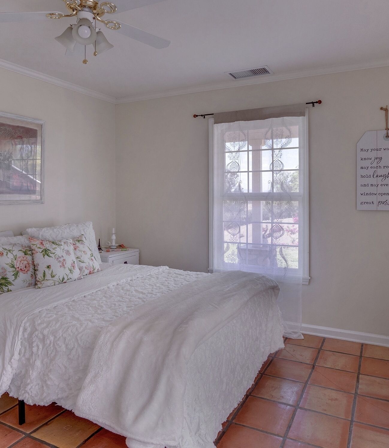 A bedroom with a large bed, a ceiling fan, a window with curtains, and a wall-mounted sign.