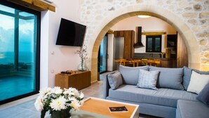 A modern living room with a grey sofa, a wooden coffee table, and a TV mounted on the wall. There is a stone archway leading to a kitchen area with wooden cabinets and a dining table.