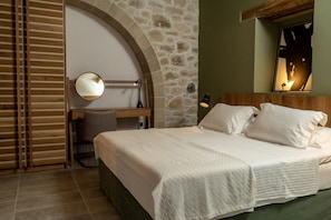 A bedroom with a stone wall, a wooden desk, a bed with white bedding, and a nightstand with lamps.