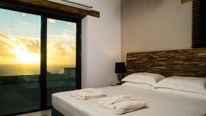 A bedroom with a large bed, a wooden headboard, a nightstand with a lamp, and a view of the sunset through a sliding door.