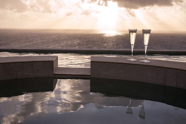 A serene pool area with two champagne flutes on a platform, overlooking a calm body of water and a sunset.