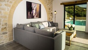 A living room with a stone wall, a grey sofa, a painting, a lamp, and a view of a swimming pool.