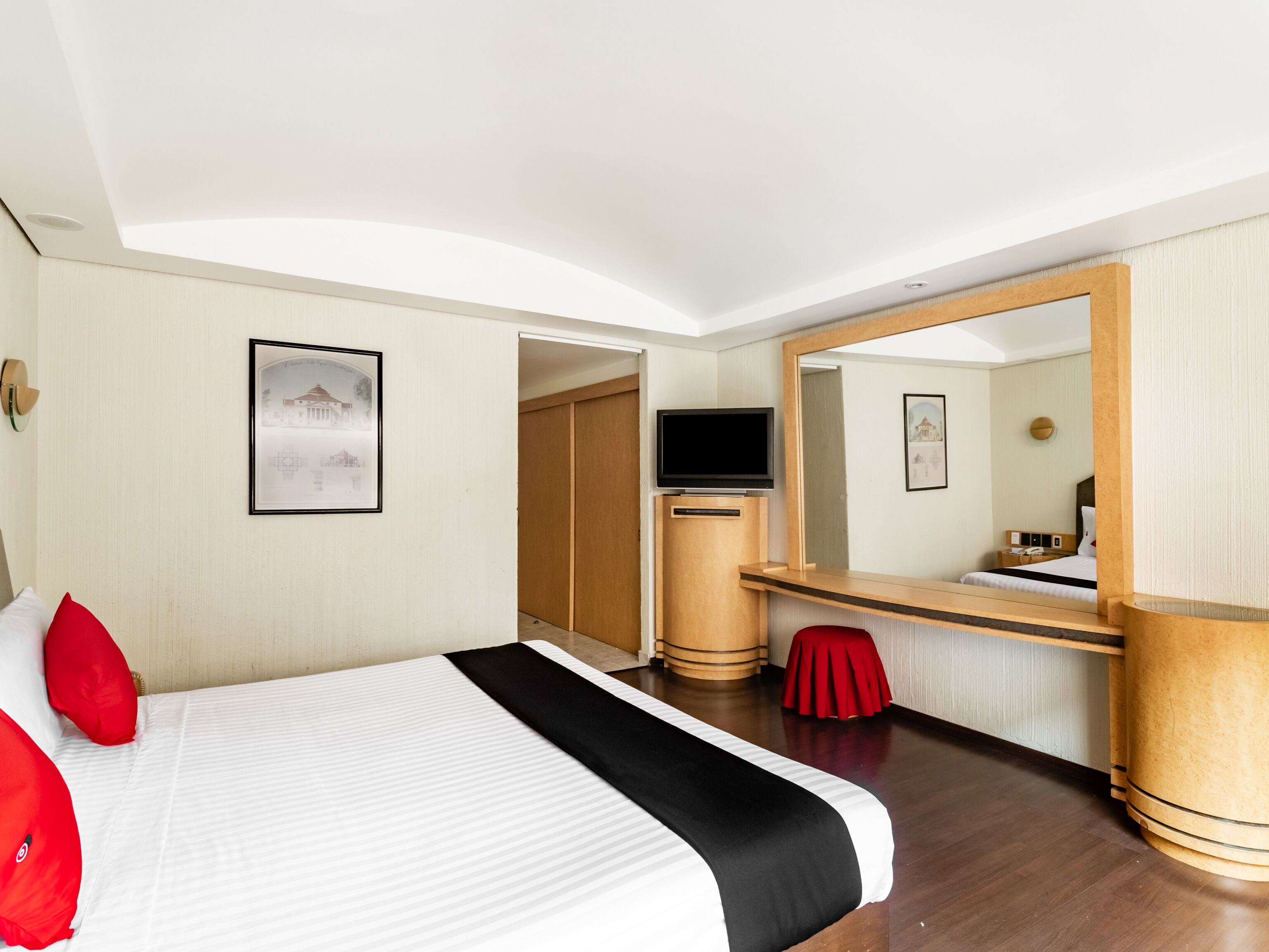 Quarto de hotel com cama, escrivaninha, televisão e espelho.