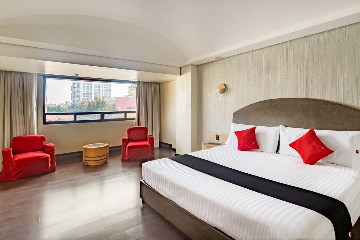 A hotel room with a large bed, two red chairs, a small wooden table, and a view of buildings through a large window.