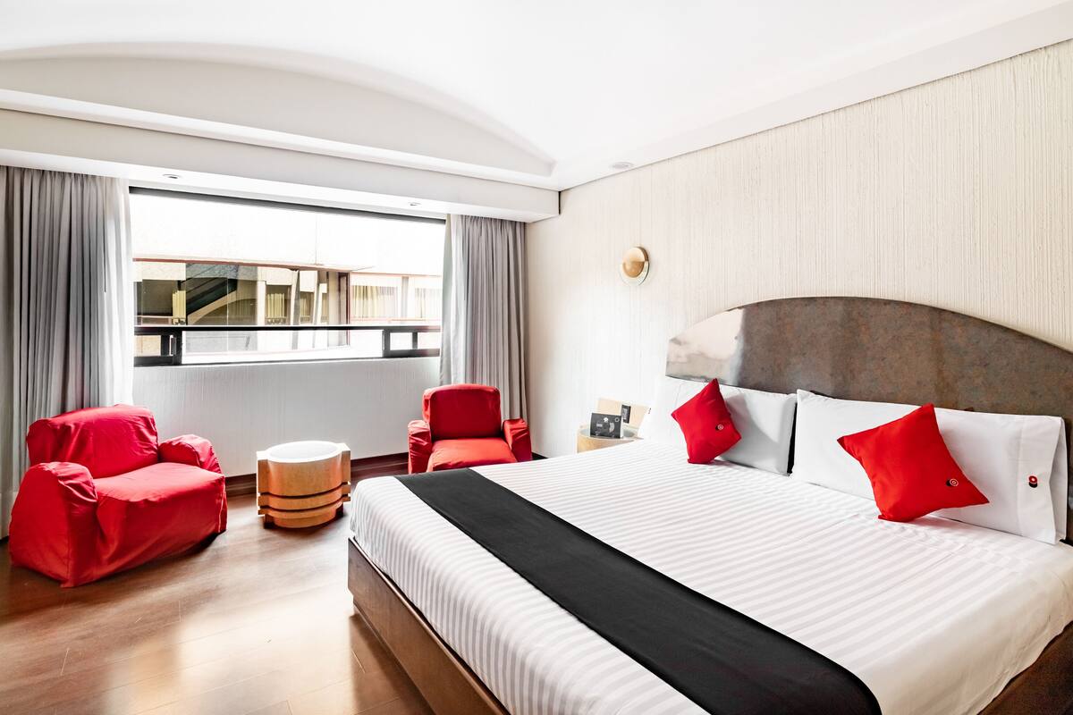 A modern hotel room with a large bed, red armchairs, a round ottoman, and a view of a building through a large window.