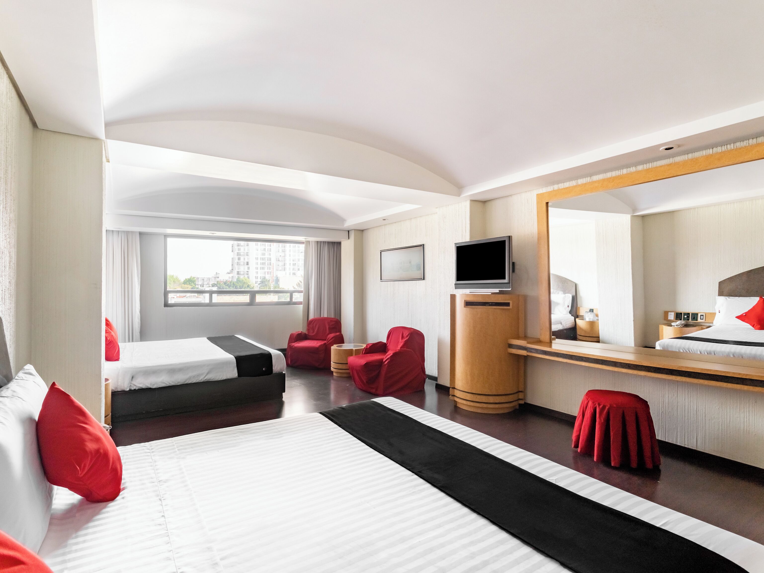 Habitación de hotel con dos camas, televisión, escritorio, espejo y ventana con vista a edificios.
