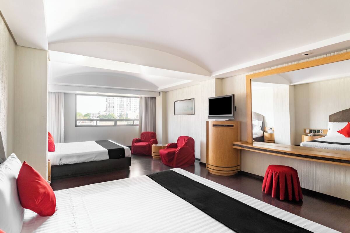 A hotel room with two beds, a TV, a desk, a mirror, and a window with a view of buildings.