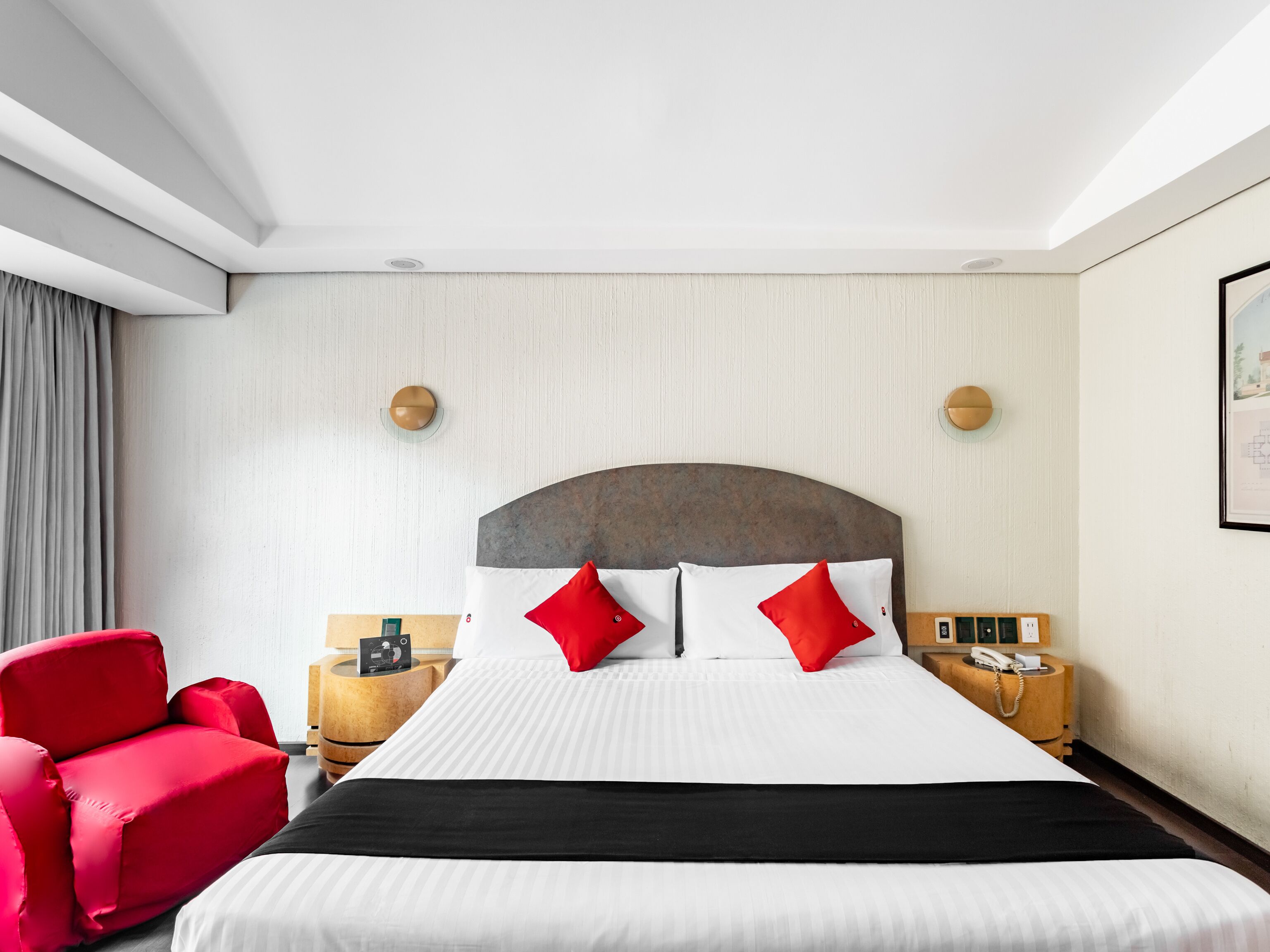 Ein Hotelzimmer mit einem großen Bett, roten und weißen Kissen, einem roten Sessel, einem Nachttisch aus Holz mit einem Telefon und einem gerahmten Bild an der Wand.