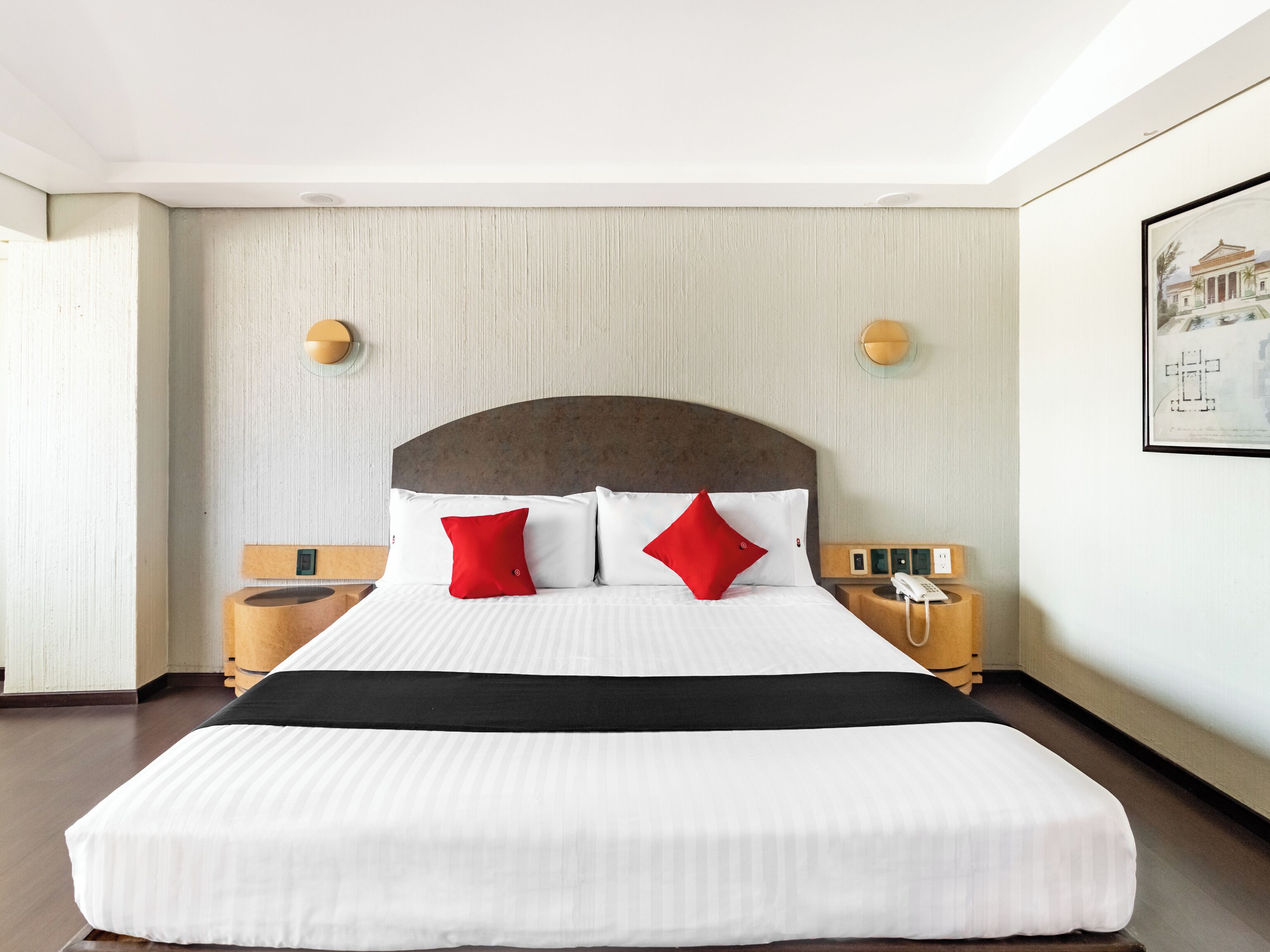 Habitación de hotel con una cama grande, dos mesitas de noche, un teléfono en la pared y un cuadro enmarcado.
