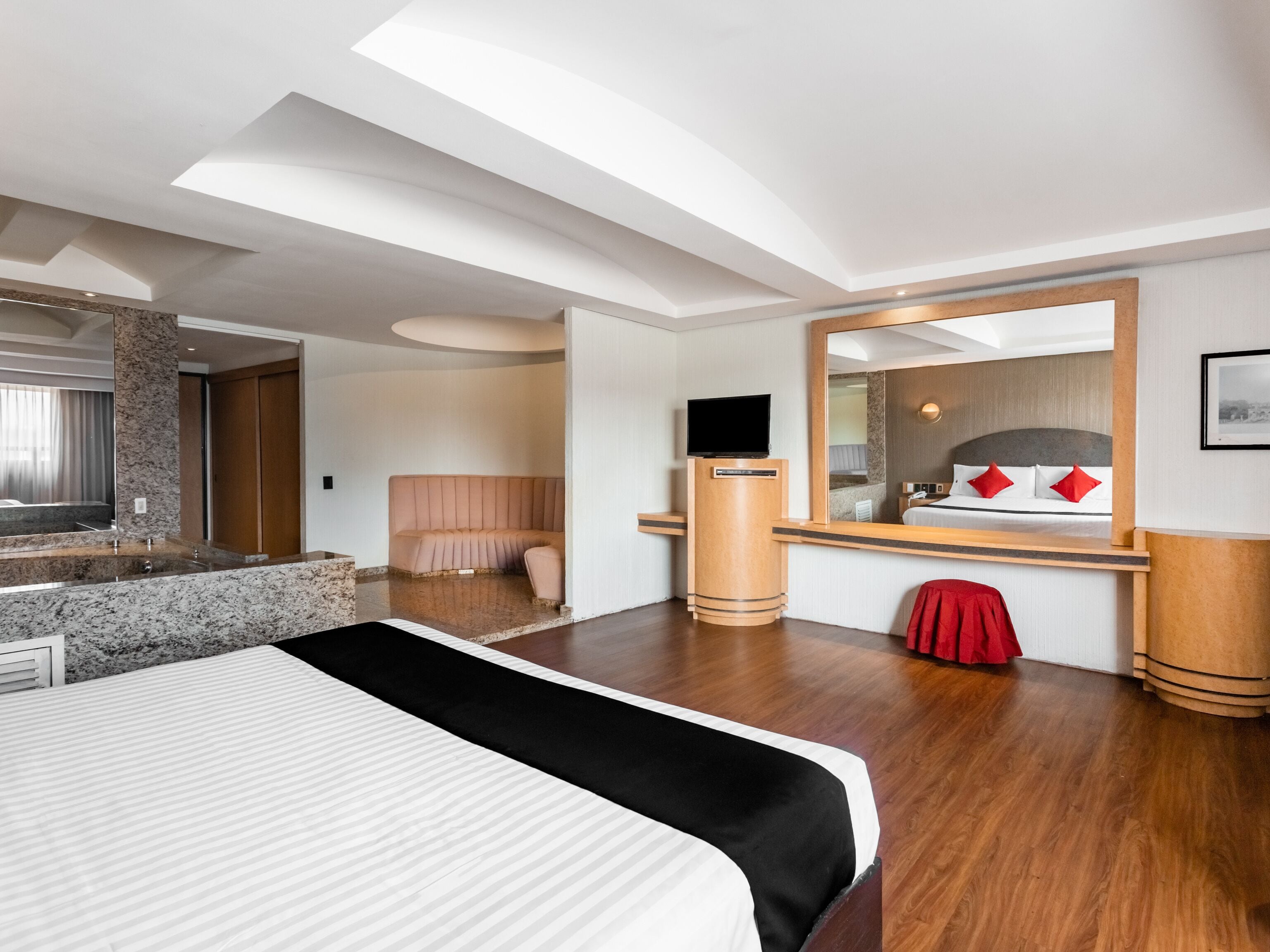 A modern hotel room with a large bed, a vanity with a mirror, a television, and a red chair.