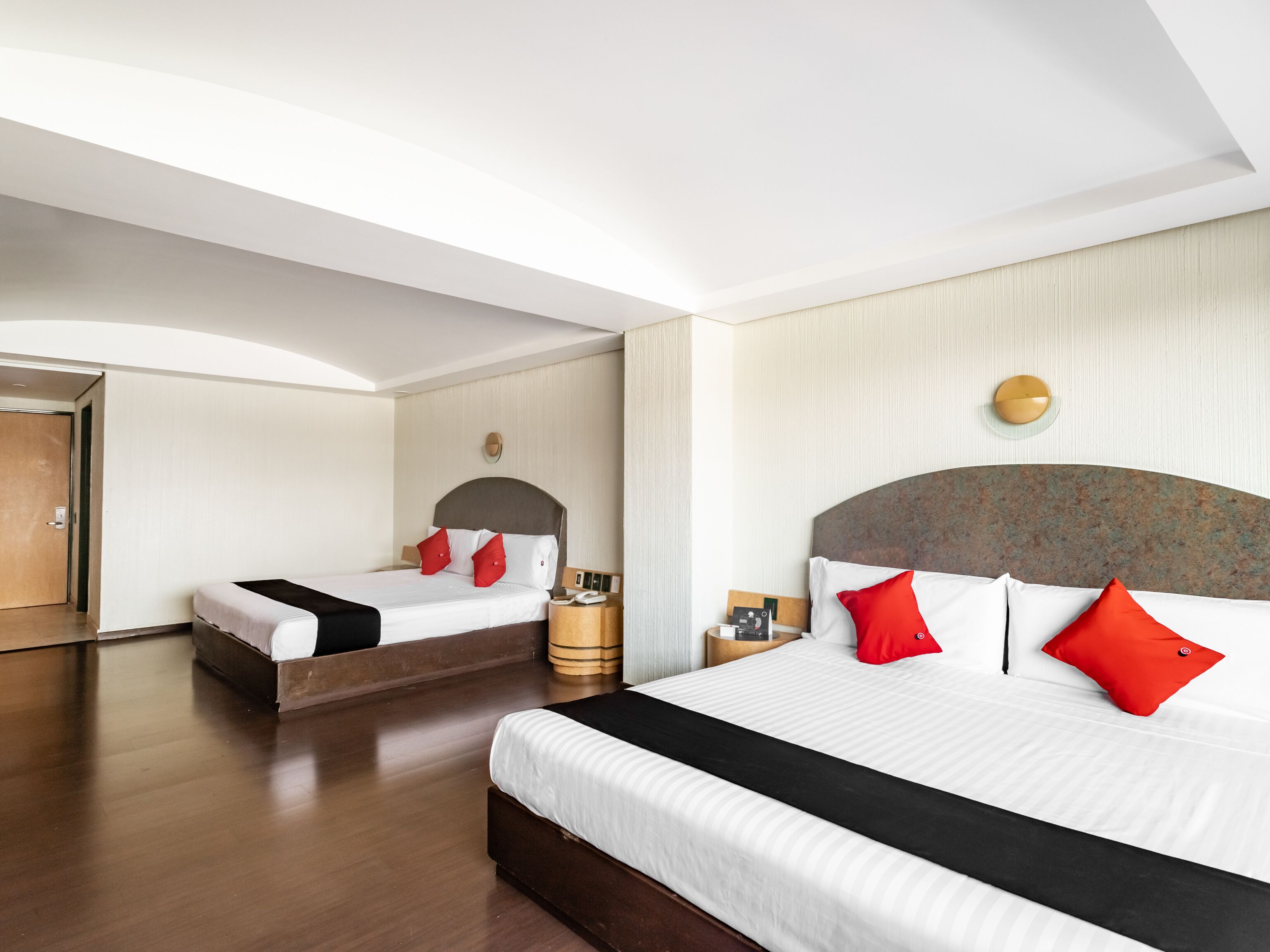 A hotel room with two beds, a wooden floor, a large mirror, and a wall-mounted light fixture.