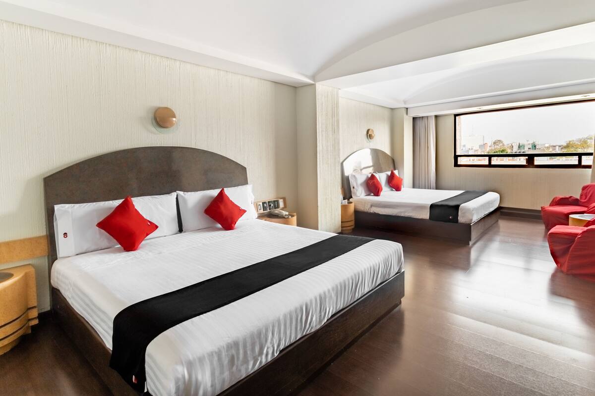 A hotel room with a large bed, a smaller bed, a red chair, and a view of the city.