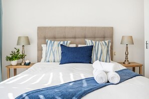 A neatly made bed with a blue blanket, two pillows, and rolled towels on a wooden nightstand.