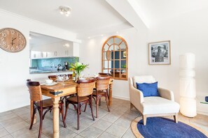 A dining area with a wooden table and chairs, a kitchen in the background, a large mirror, and a framed picture on the wall.