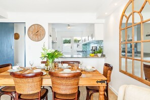 A dining area with a wooden table set for four, surrounded by wicker chairs. A large round wall clock and a mirror are visible.