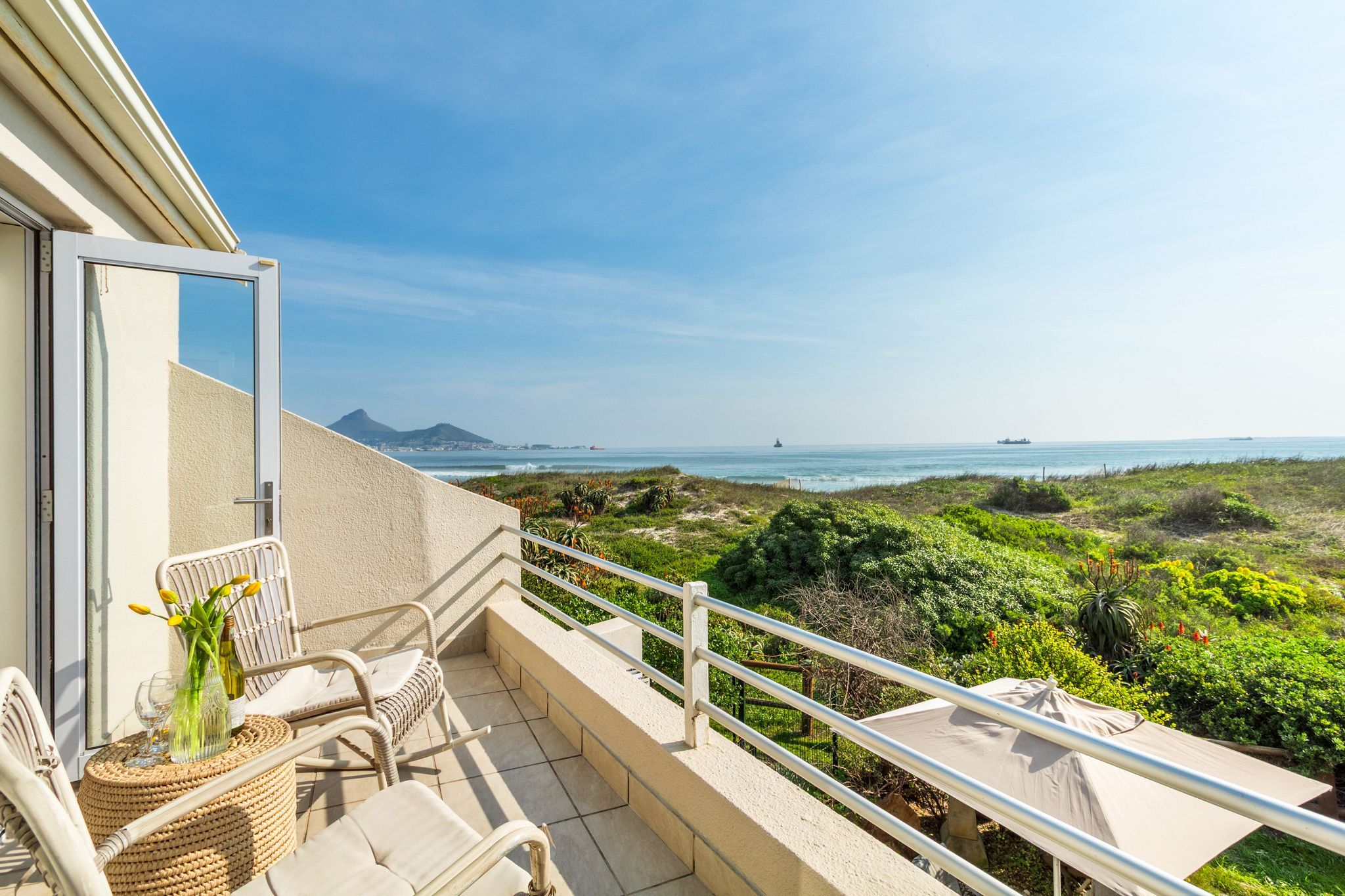 A balcony with wicker chairs, a small table with a vase, and a view of the sea and greenery.