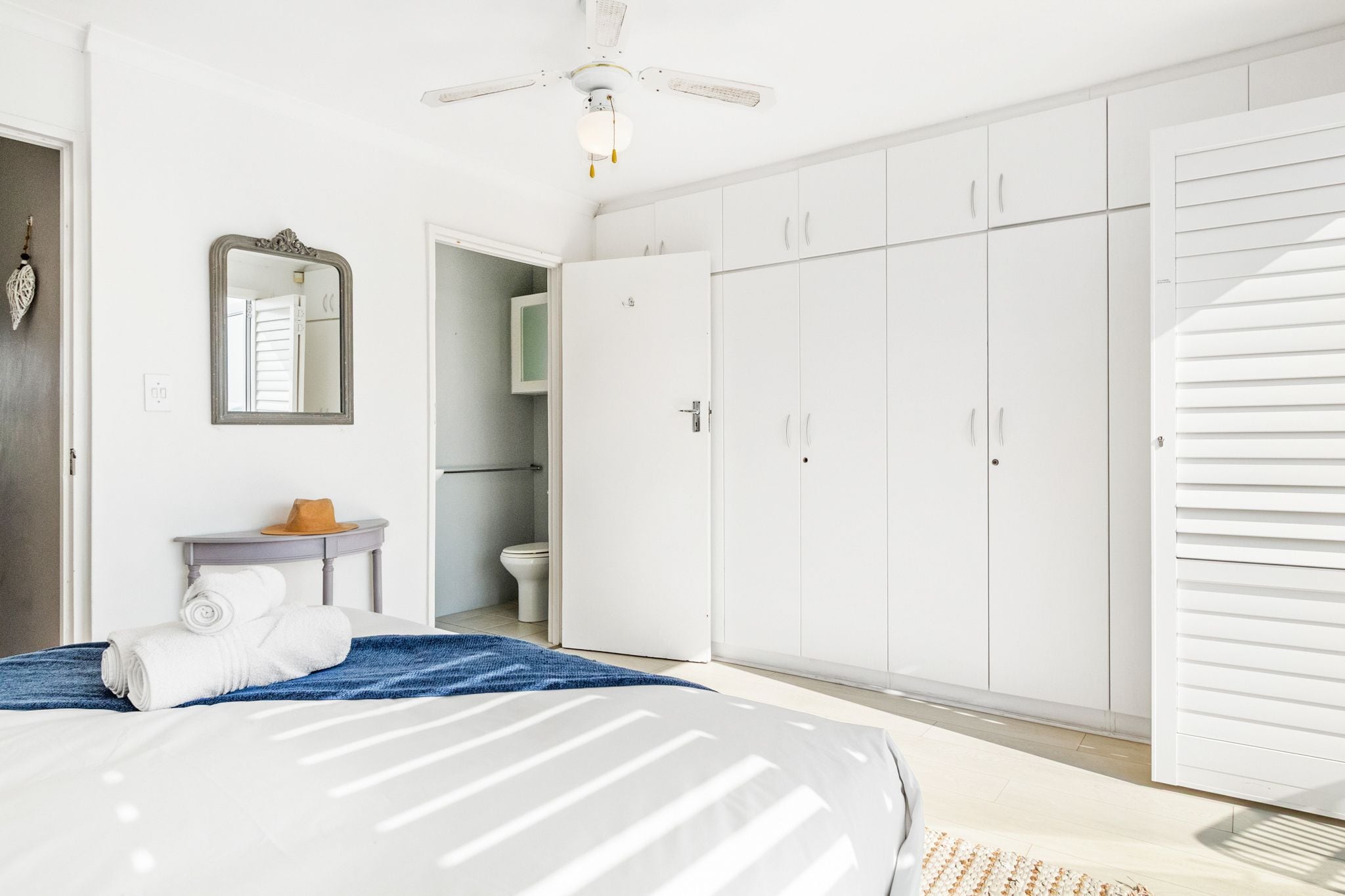 A bedroom with a bed, a wardrobe, a mirror, a small table with a hat, and a ceiling fan.