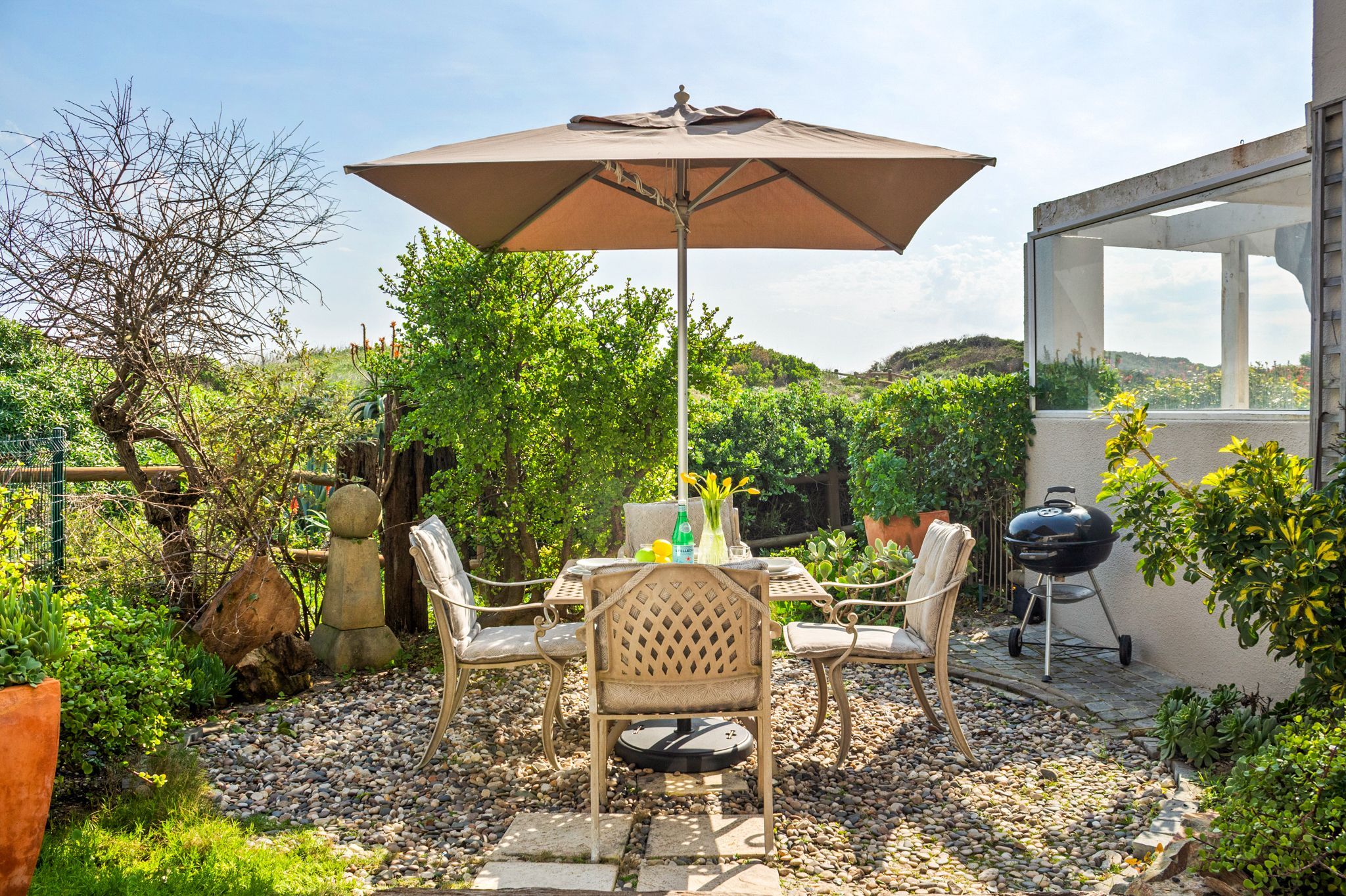 An outdoor patio with a table and chairs under a large umbrella, surrounded by greenery and a barbecue grill.