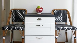 Una habitación con dos sillones de mimbre con cojines de estampado blanco y negro, un aparador blanco con cajones y una repisa de madera sobre el aparador que sostiene dos frascos de vidrio con flores secas y un letrero enmarcado que dice 'Amour sans fin'.